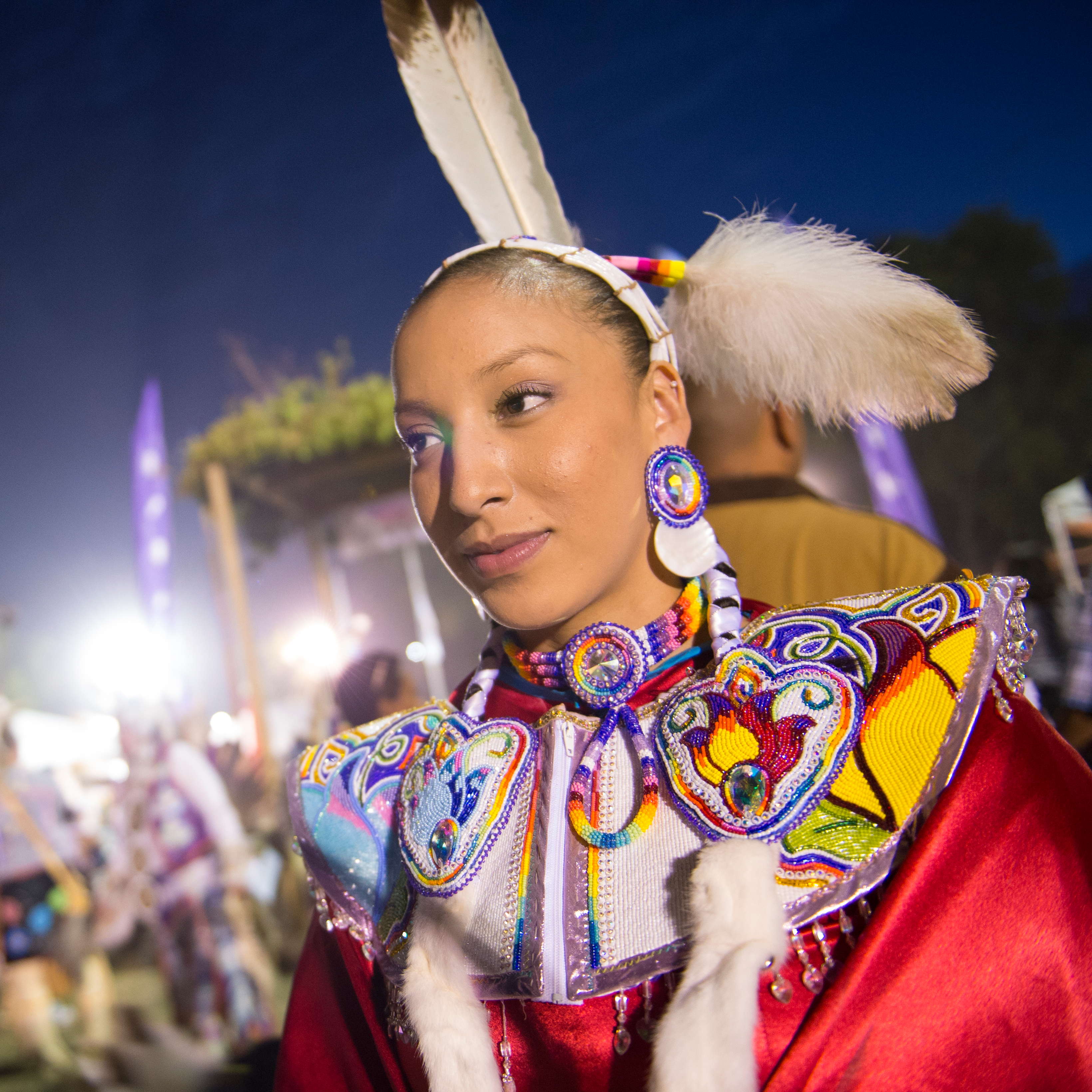 Photo Courtesy of Rebecca Comfort A woman dressed in red and rainbow regalia with many beaded floral pieces and beaded collar, stands proud in the foreground of a croud blurry behind her. She has a long beaded headband with feathers on her head, her hair braided tightly with two wrapped braids down her front.